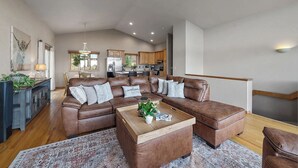 A spacious living room with a brown leather sectional sofa, a wooden coffee table, and a patterned rug. A kitchen is visible in the background with wooden cabinets and a refrigerator. - Luxury Evergreen Retreat · Fireplace · Mountain Views · 45 min Denver  (Evergreen)