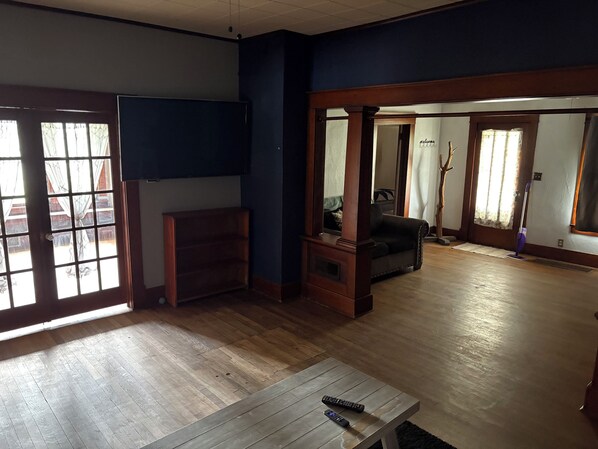 A spacious living room with wooden flooring, a flat-screen TV mounted on the wall, a dark leather sofa, and a coffee table with remotes.