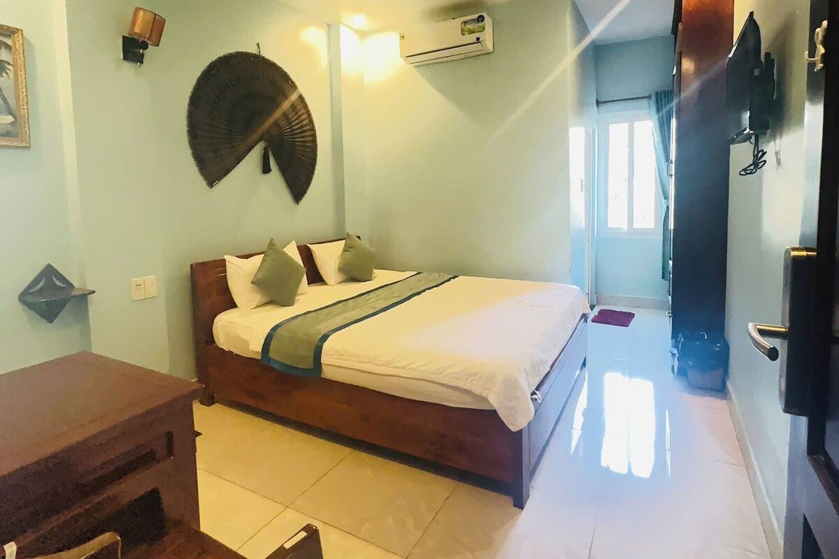 A bedroom with a bed, a desk, and a chair. There is a window with curtains, an air conditioning unit, and a wall-mounted TV.