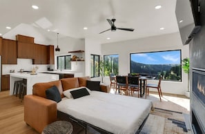Cozinha moderna com armários de madeira e ilha central, área de estar acolhedora com sofá e cadeiras, e uma janela grande com vista para o exterior.