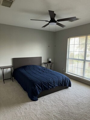 A bedroom with a bed, a ceiling fan, a bedside table, a lamp, and a window with blinds.