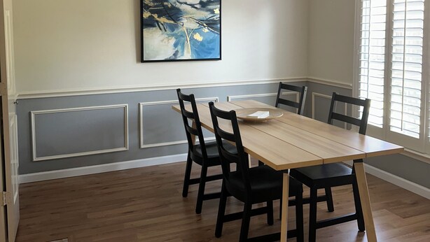 Un comedor con mesa y sillas de madera, una lámpara de techo, un ventanal con persianas y un cuadro abstracto colgado en la pared.