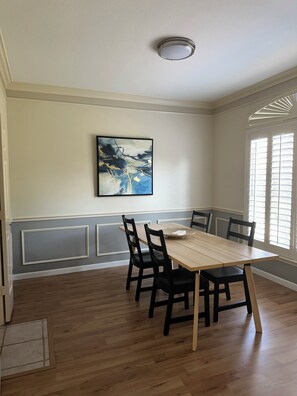 A dining area with a wooden table and chairs, a ceiling light, a window with blinds, and a wall-mounted abstract painting.