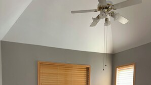 Un dormitorio con una cama, dos mesitas de noche, un ventilador de techo y dos ventanas con persianas.