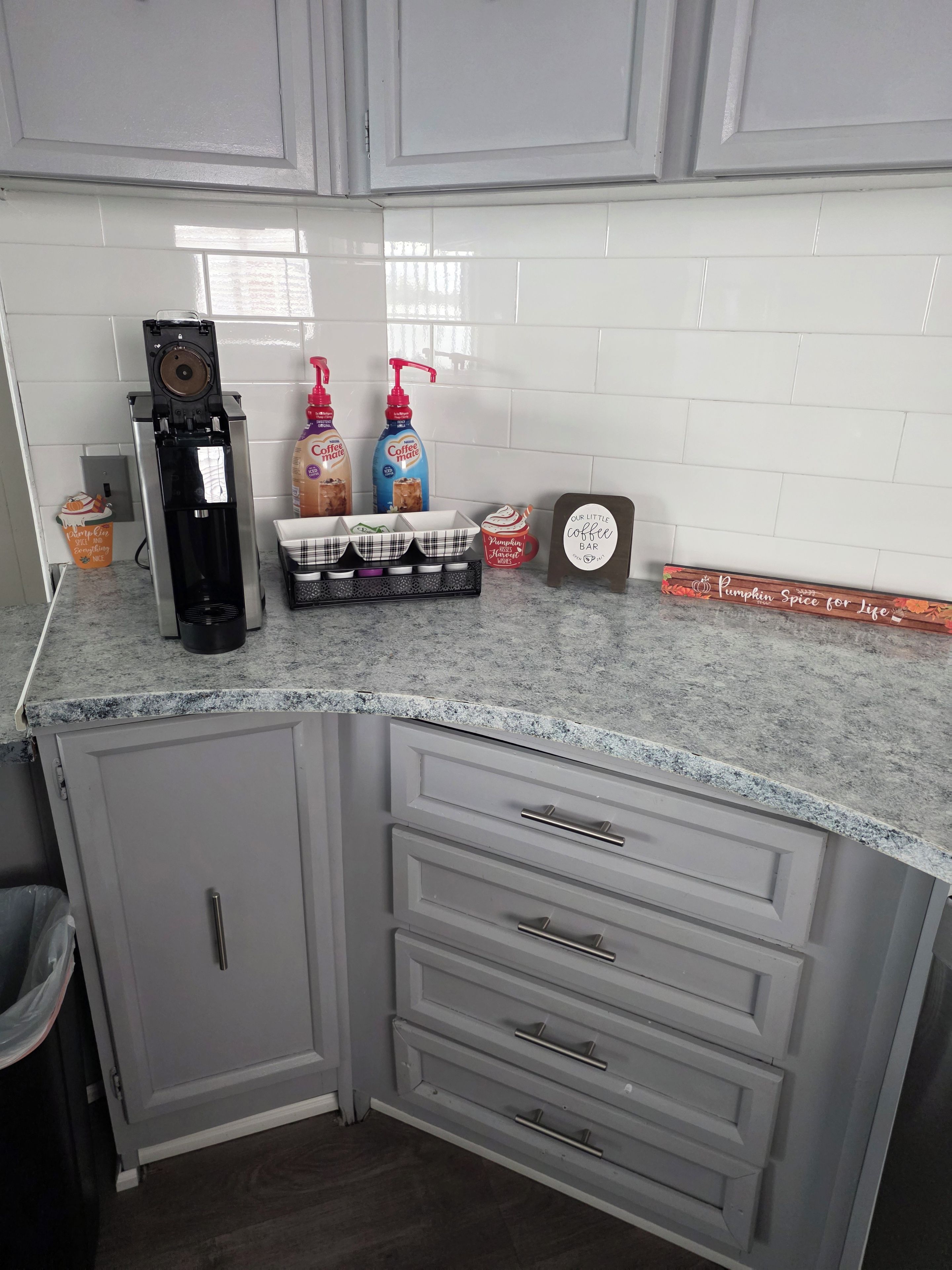 A kitchen with a coffee machine, two bottles of coffee, a pumpkin spice mug, a coffee bar sign, and a decorative sign.