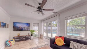 A spacious living room with a ceiling fan, a large TV, a dark-colored sofa with decorative pillows, a wooden coffee table, and a colorful basket.