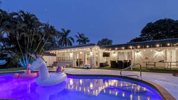 A swimming pool with a unicorn float, a covered patio area, and palm trees in the background.