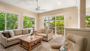 A spacious living room with a beige sofa, a wooden coffee table, and a ceiling fan. Large windows provide a view of lush greenery outside.