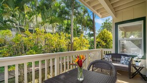 A covered outdoor seating area with wicker furniture and a white railing, surrounded by lush tropical vegetation.