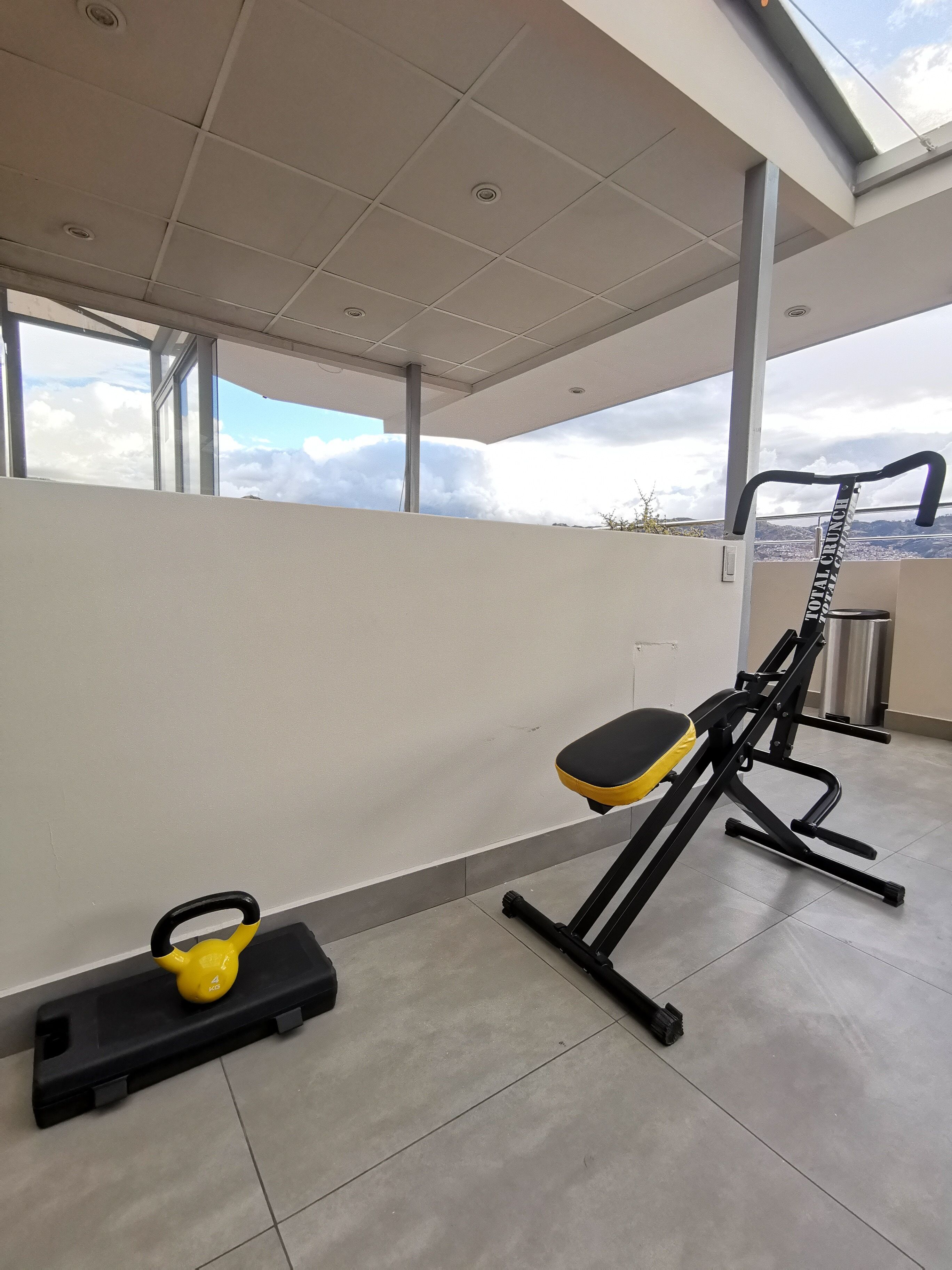 A gym with exercise equipment, including a bench and a weight machine, with a view of the sky and clouds through large windows.
