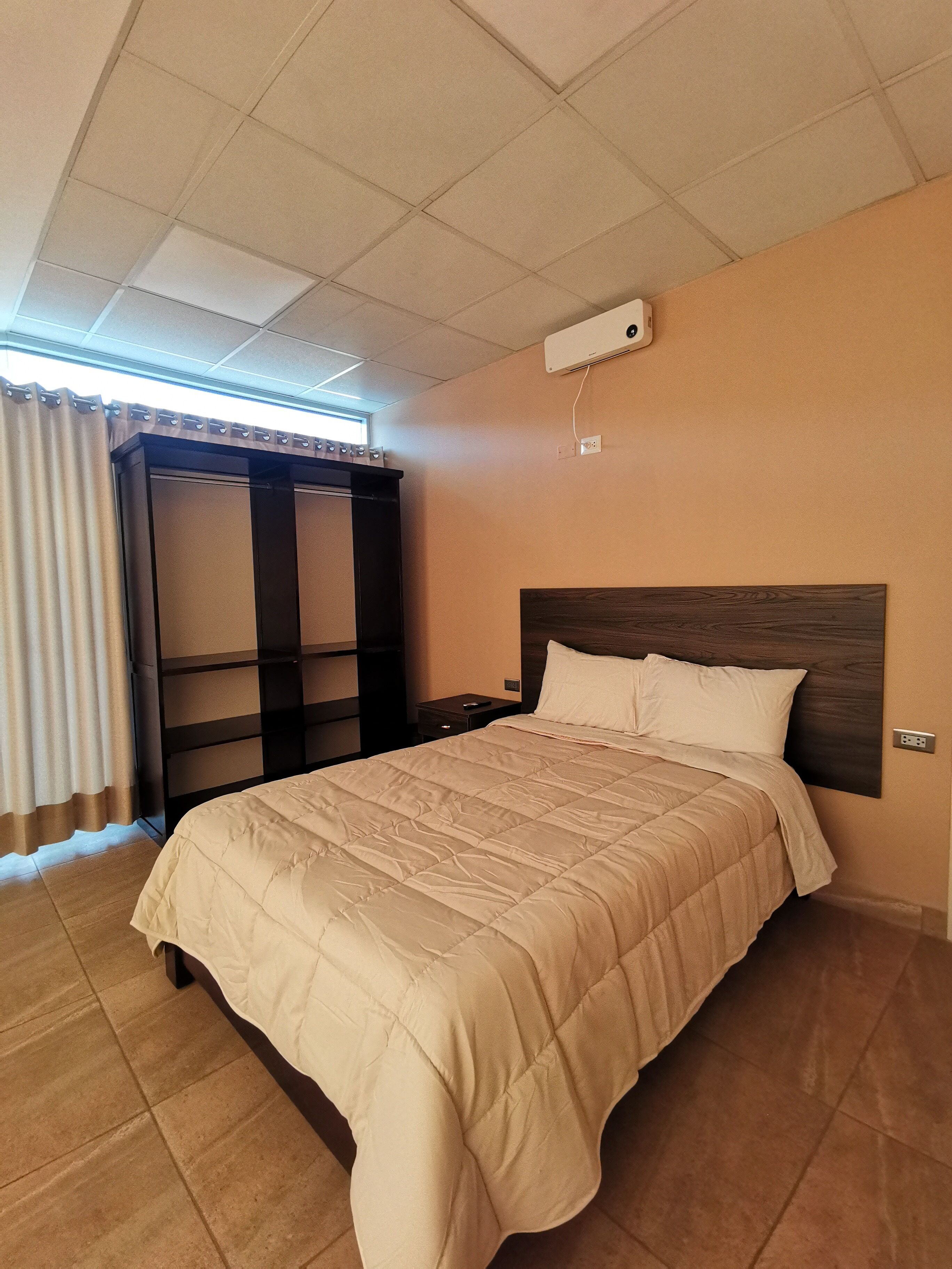 Hotelkamer met een bed, nachtkastje, wandairco en een raam met gordijnen.
