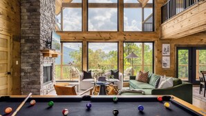A pool table in a room with a stone fireplace, large windows, and a view of the outdoors. - Chara Vista Cabin: A Smoky Mountain Retreat (Sevierville)