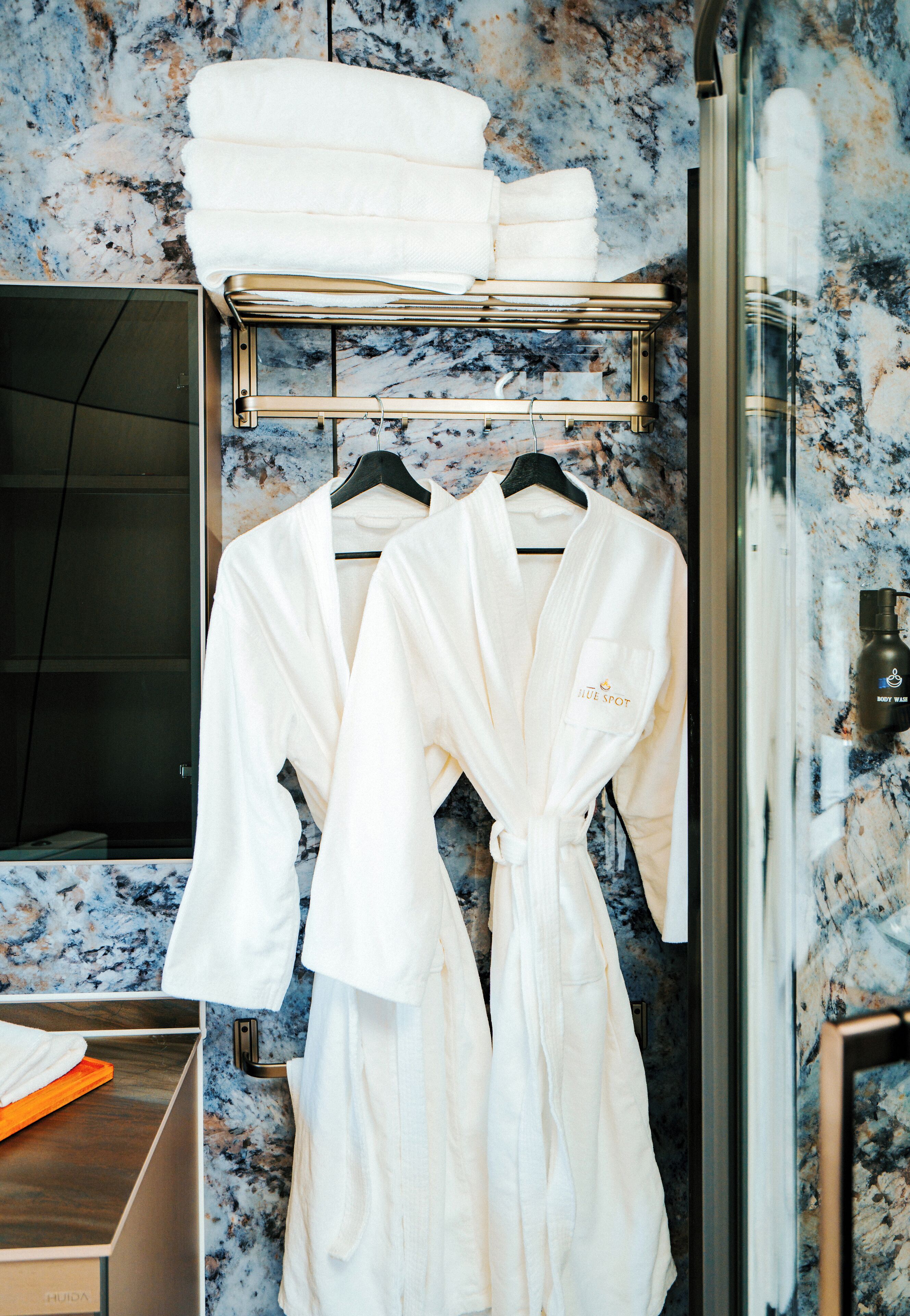 A bathroom with marble walls, two white robes hanging on a rack, and a glass shower enclosure.