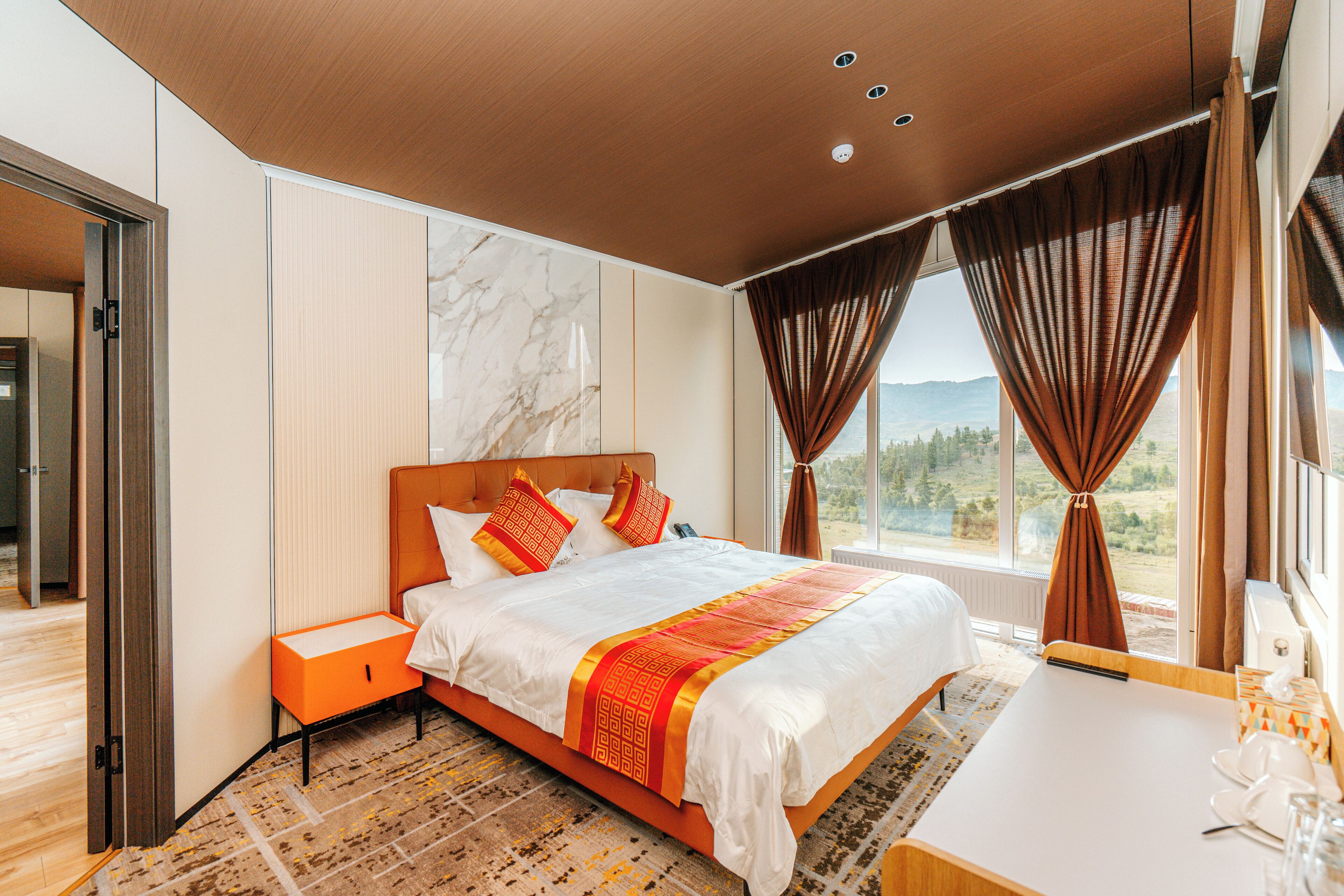 A modern hotel room with a large bed, a bedside table, a small desk, and a view of the outdoors.