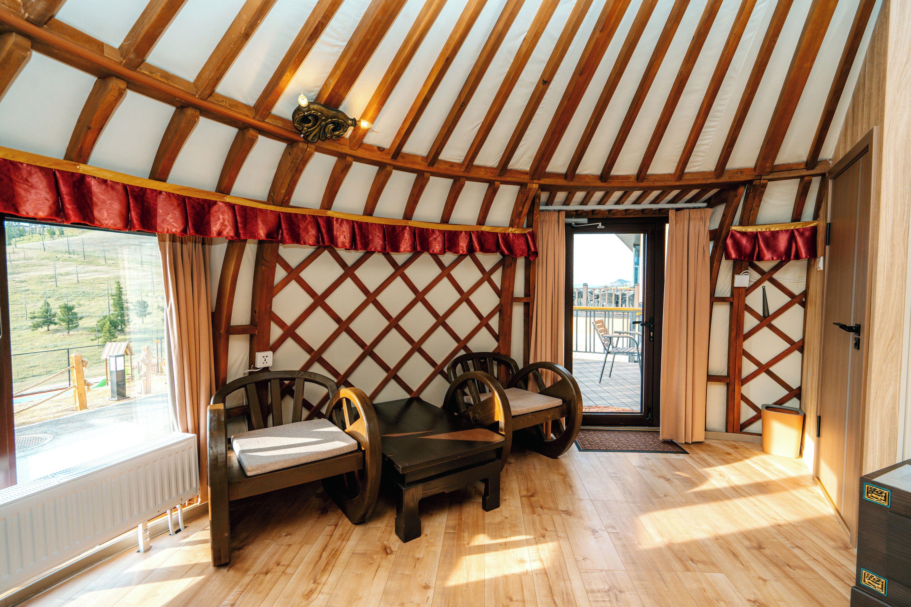 A yurt interior with a wooden floor, a table with chairs, a view through the window, and a door leading outside.