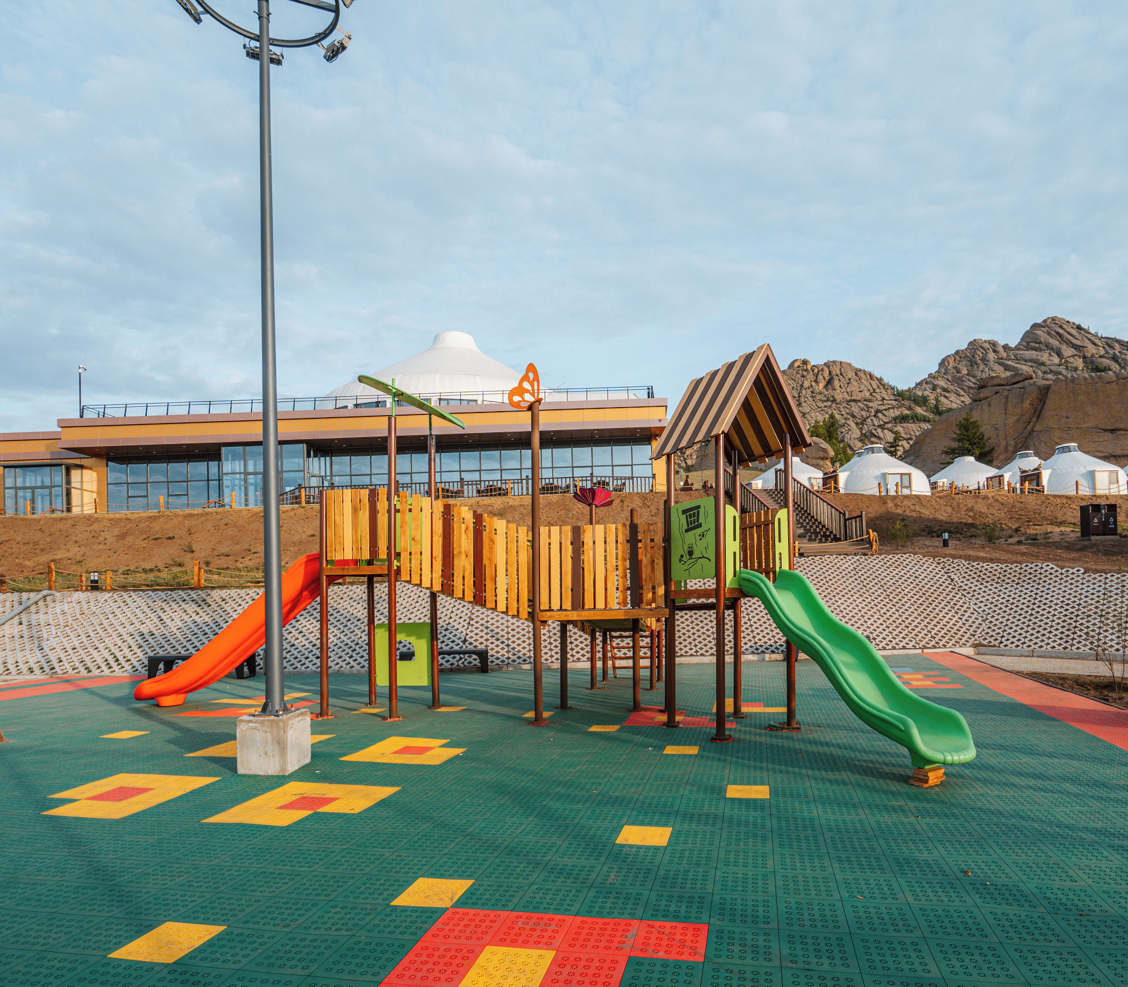 Un parque infantil con toboganes y estructuras para escalar, frente a un edificio y terreno rocoso.