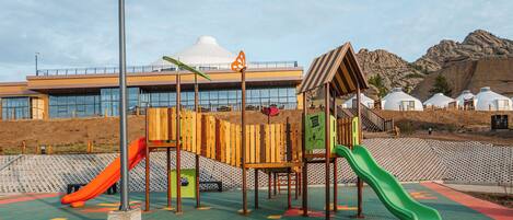 Un parque infantil con toboganes y estructuras para escalar, frente a un edificio y terreno rocoso.