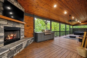 A covered outdoor area with a stone fireplace, a grill, and a seating area with a view of trees.