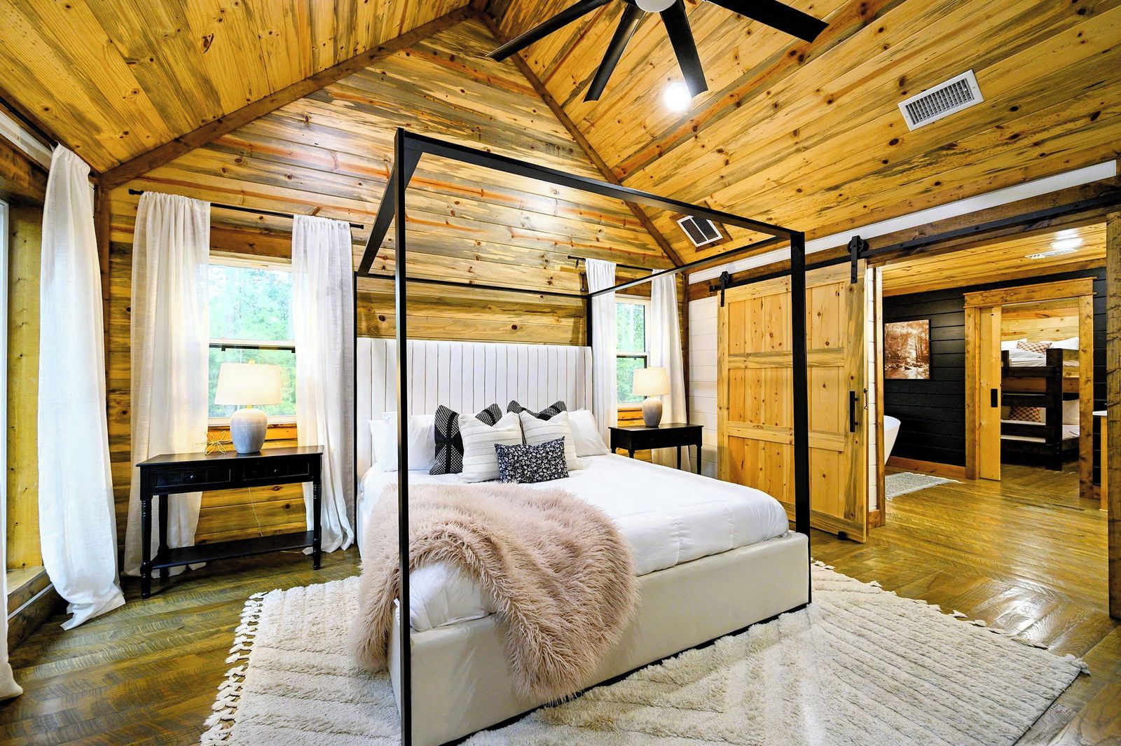 A bedroom with a four-poster bed, a wooden ceiling, and a large window with curtains.