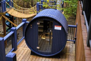 A cylindrical wooden structure with a door, surrounded by a wooden deck and a rope bridge.