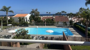 Una piscina con lettini e ombrelloni, circondata da palme e edifici con tetti in tegole rosse.