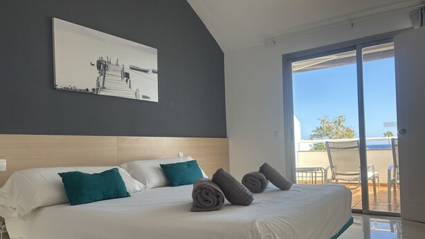 Una moderna camera d'albergo con un ampio letto, biancheria da letto color petrolio e vista sull'esterno tramite una porta scorrevole in vetro.