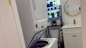 Een kleine wasruimte met een gestapelde wasmachine en droger, een klein plankje met toiletartikelen en een groen kleed.