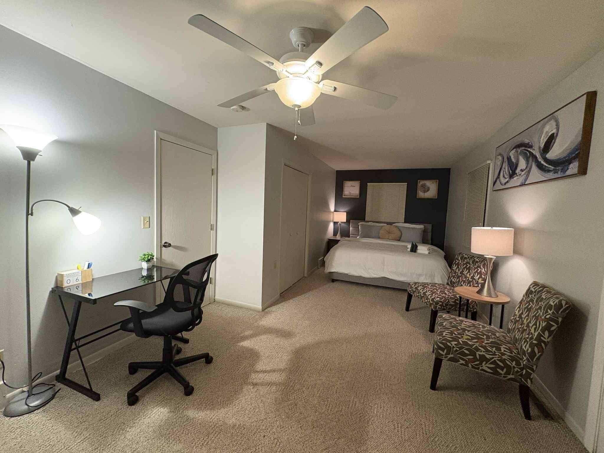 A hotel room with a bed, desk, office chair, two patterned chairs, a ceiling fan, and a lamp.