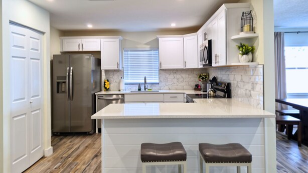 Una cocina moderna con armarios blancos, un refrigerador de acero inoxidable, un protector contra salpicaduras de mármol y una isla central con dos taburetes.