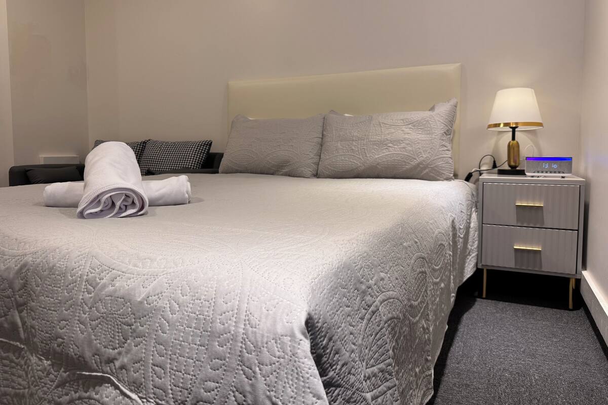 Cama bem arrumada com toalha dobrada, abajur e relógio digital em uma mesa de cabeceira.