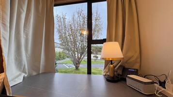 Habitación de hotel con vistas a un árbol y un estacionamiento a través de la ventana. Hay una lámpara en la mesa y un cargador de teléfono a un lado.