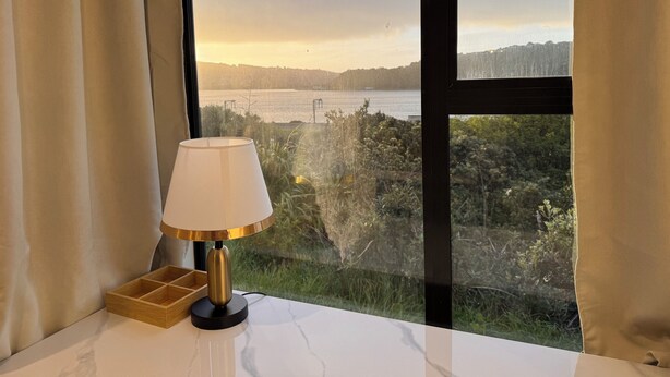 A room with a view of a body of water and greenery, a table with a lamp, and a wooden tray. - Harbour View Lodge (Porirua)