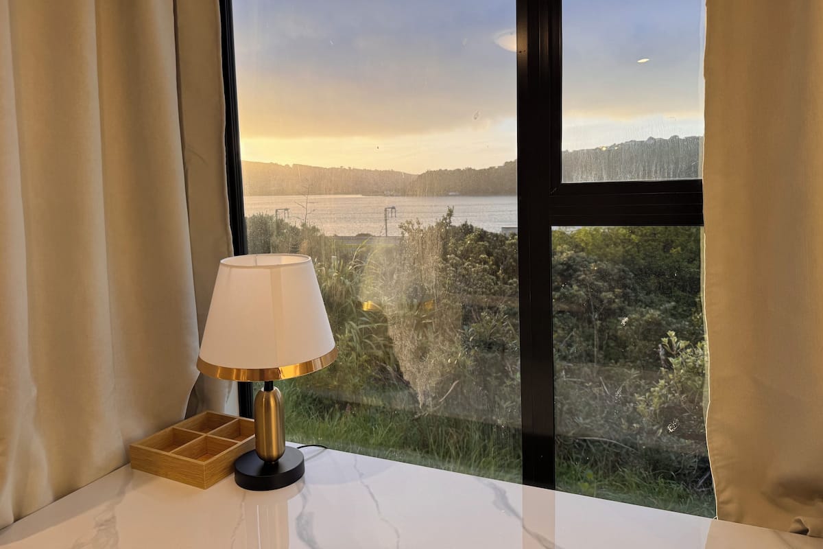 Quarto com vista para a água e vegetação, uma mesa com abajur e um tabuleiro de madeira.