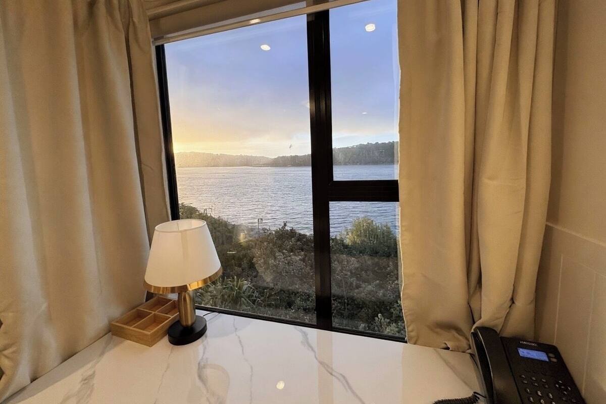 Quarto com uma grande janela com vista para um corpo d'água, uma luminária sobre uma mesa de mármore e um telefone no lado direito.