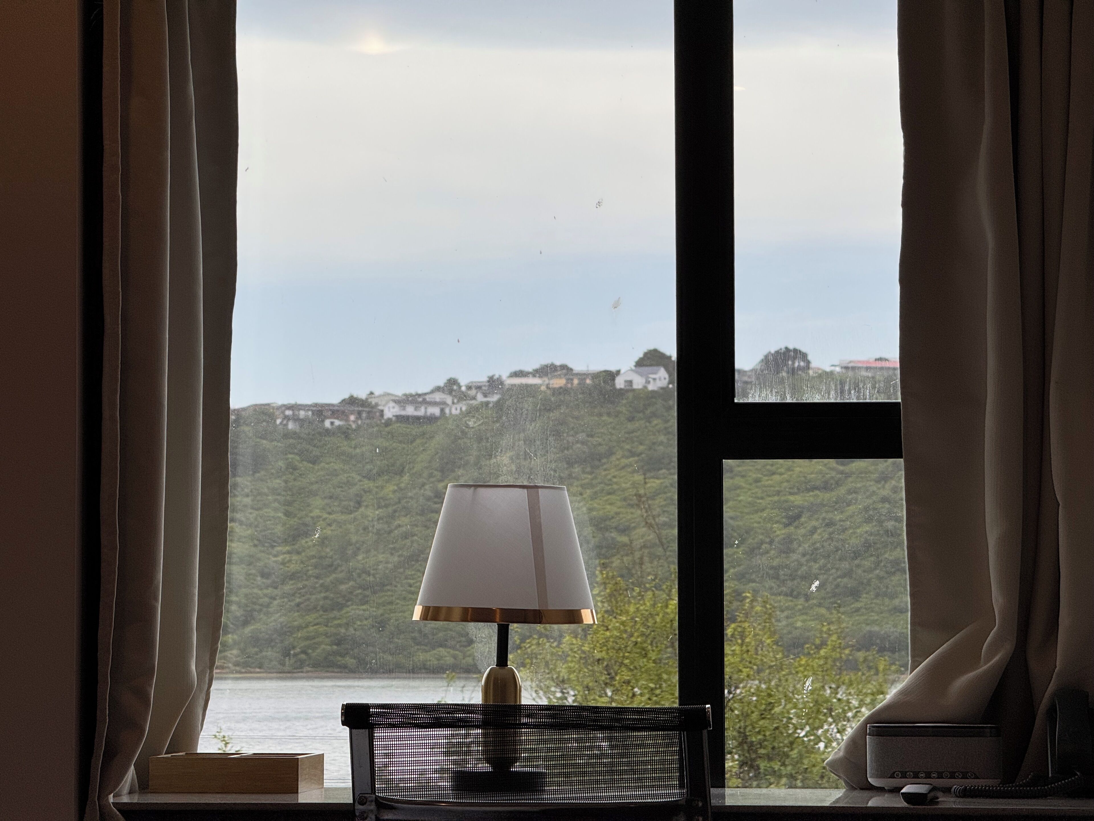 A room with a view of a hillside, a lamp on a table, and a remote control on a windowsill.