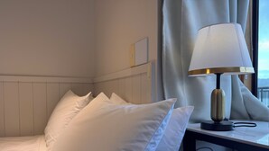 A neatly made bed with white linens and pillows, a bedside table with a lamp, and a window with curtains. - Harbour View Lodge (Porirua)