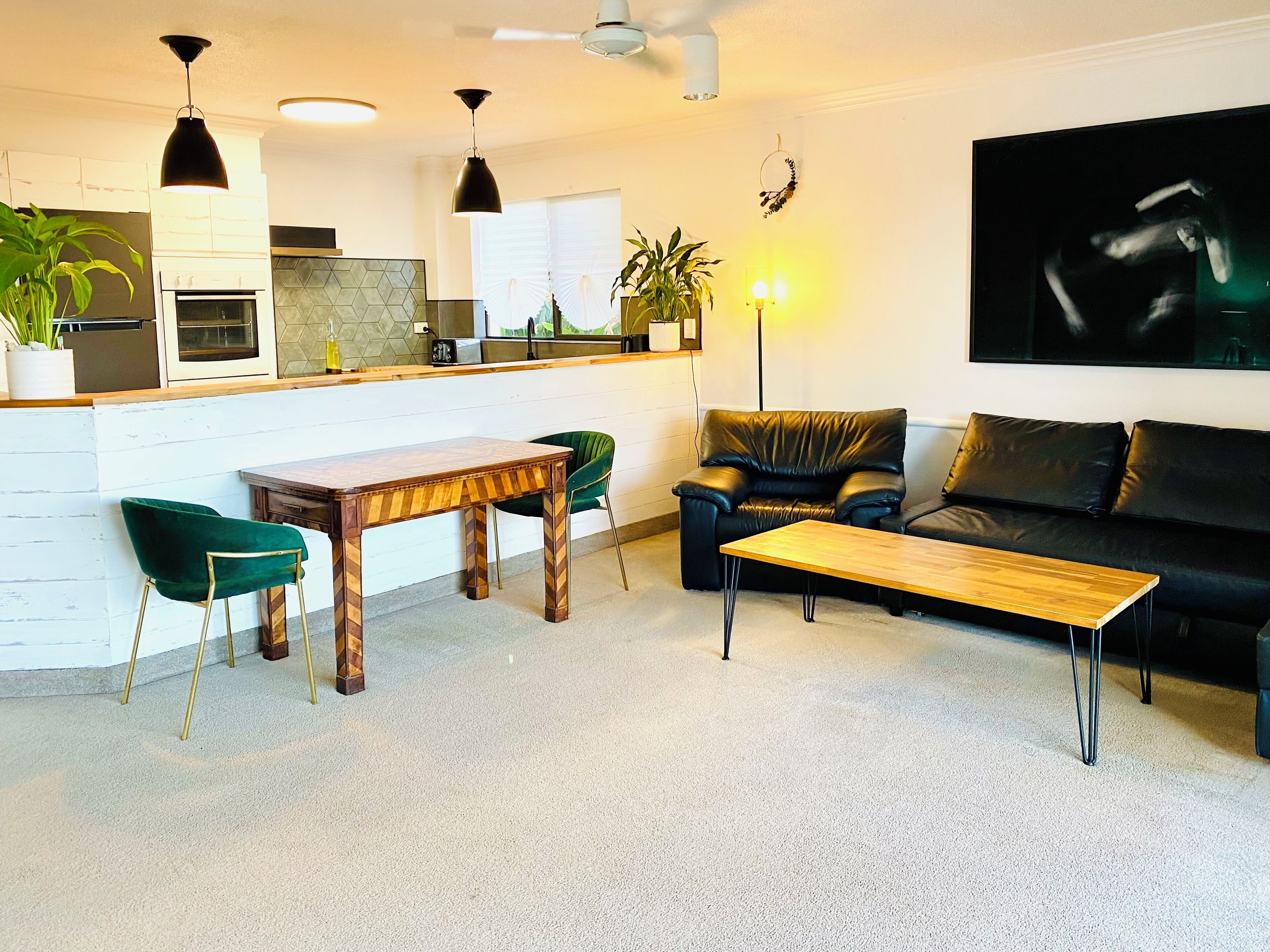 A modern living room with a wooden coffee table, a green velvet armchair, a black leather sofa, and a kitchen area with white cabinets and a black countertop.