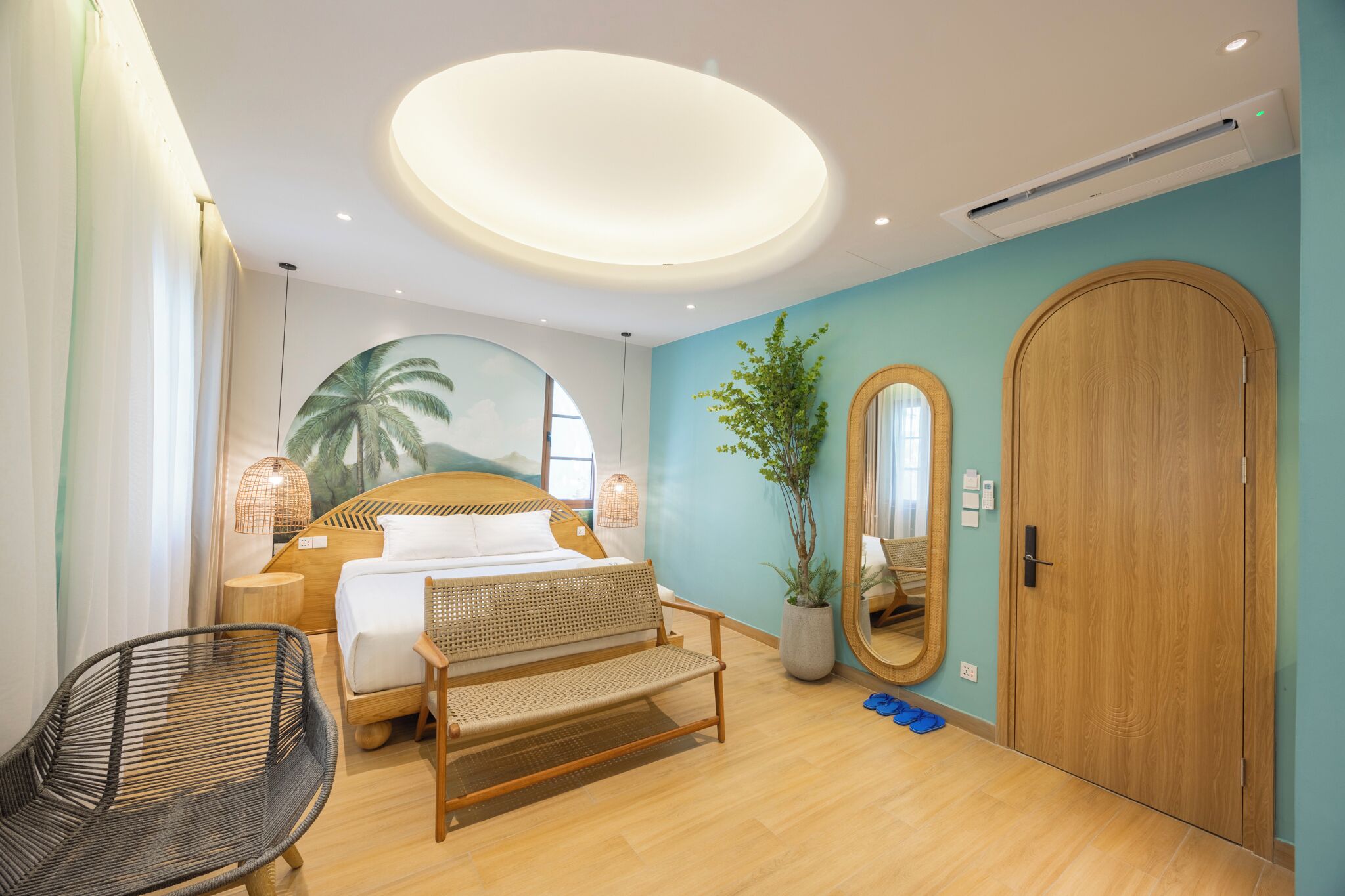 A modern hotel room with a bed, a wicker chair, a wooden wardrobe, a large mirror, and a potted plant.