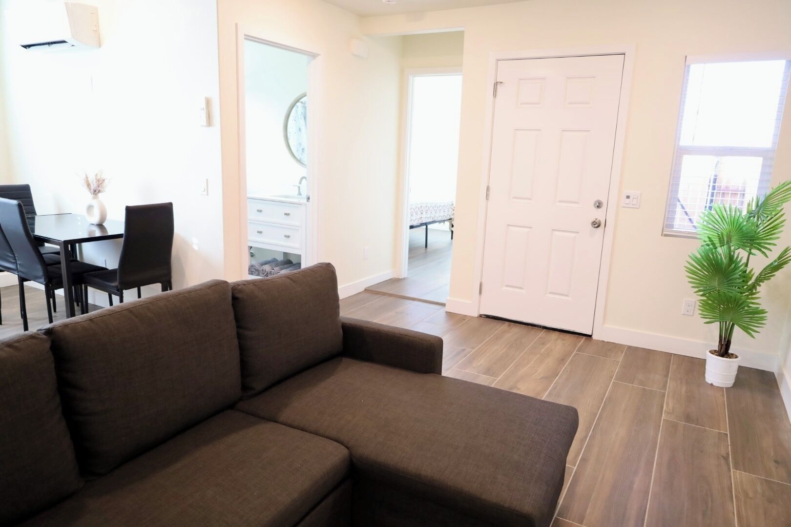 A modern living room with a brown sofa, a dining table with chairs, a white door, and a potted plant.