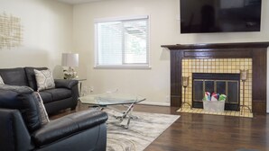 Ein Wohnzimmer mit einem schwarzen Ledersofa, einem Glastisch, einem Kamin mit einer Zeitschriftkorb und einem flachbildfernseher an der Wand.