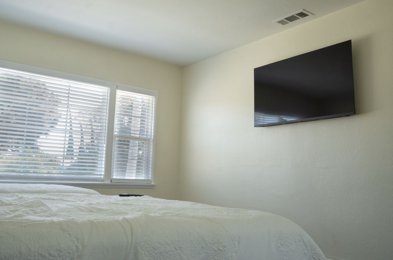 Une chambre à coucher avec un lit, deux fenêtres équipées de stores et un téléviseur à écran plat fixé au mur.