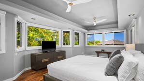 Een slaapkamer met een groot bed, een televisie op een dressoir en meerdere ramen met uitzicht op groen en een veraf gelegen landschap.