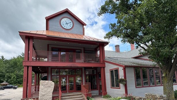 A two-story building with a clock on the upper level, a sign reading "Hospitality Building," and a stone bench in front. - Attitash Room: Pool, Hot Tub, Storyland, Foliage (Bartlett)