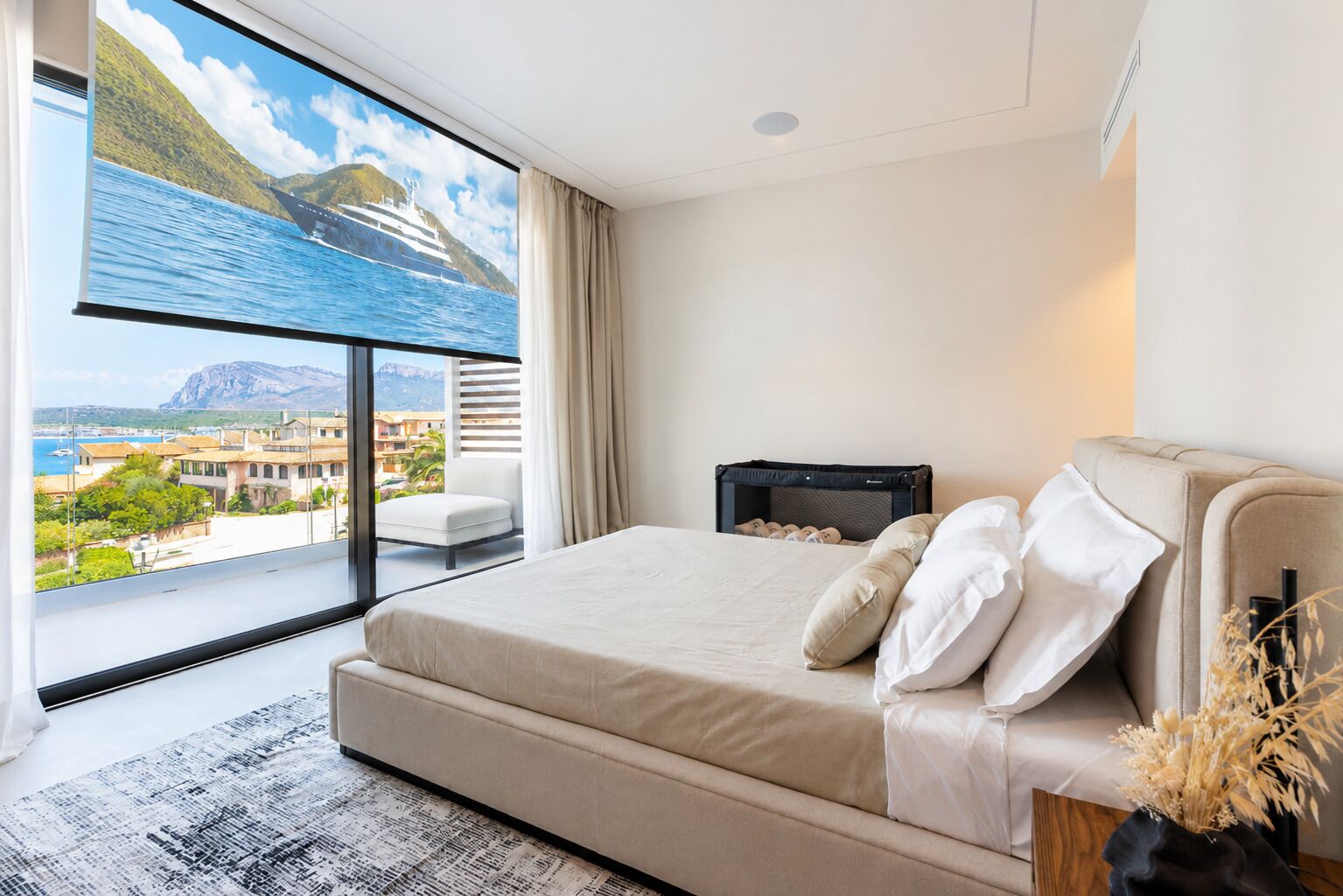 Ein modernes Schlafzimmer mit großem Fenster, einem Bett mit weißer Bettwäsche, einem kleinen Nachttisch und Blick auf eine Küstenlandschaft.
