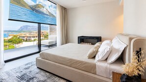 A modern bedroom with a large window, a bed with white bedding, a small bedside table, and a view of a coastal landscape.