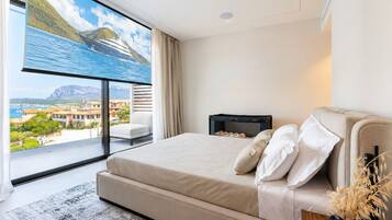 Ein modernes Schlafzimmer mit großem Fenster, einem Bett mit weißer Bettwäsche, einem kleinen Nachttisch und Blick auf eine Küstenlandschaft.