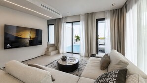 A modern living room with a flat-screen TV, a sofa, a coffee table, and a view of a pool through large windows.