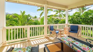 Een overdekt balkon met rieten meubilair, een kleine tafel en uitzicht op weelderige begroeiing en palmbomen.