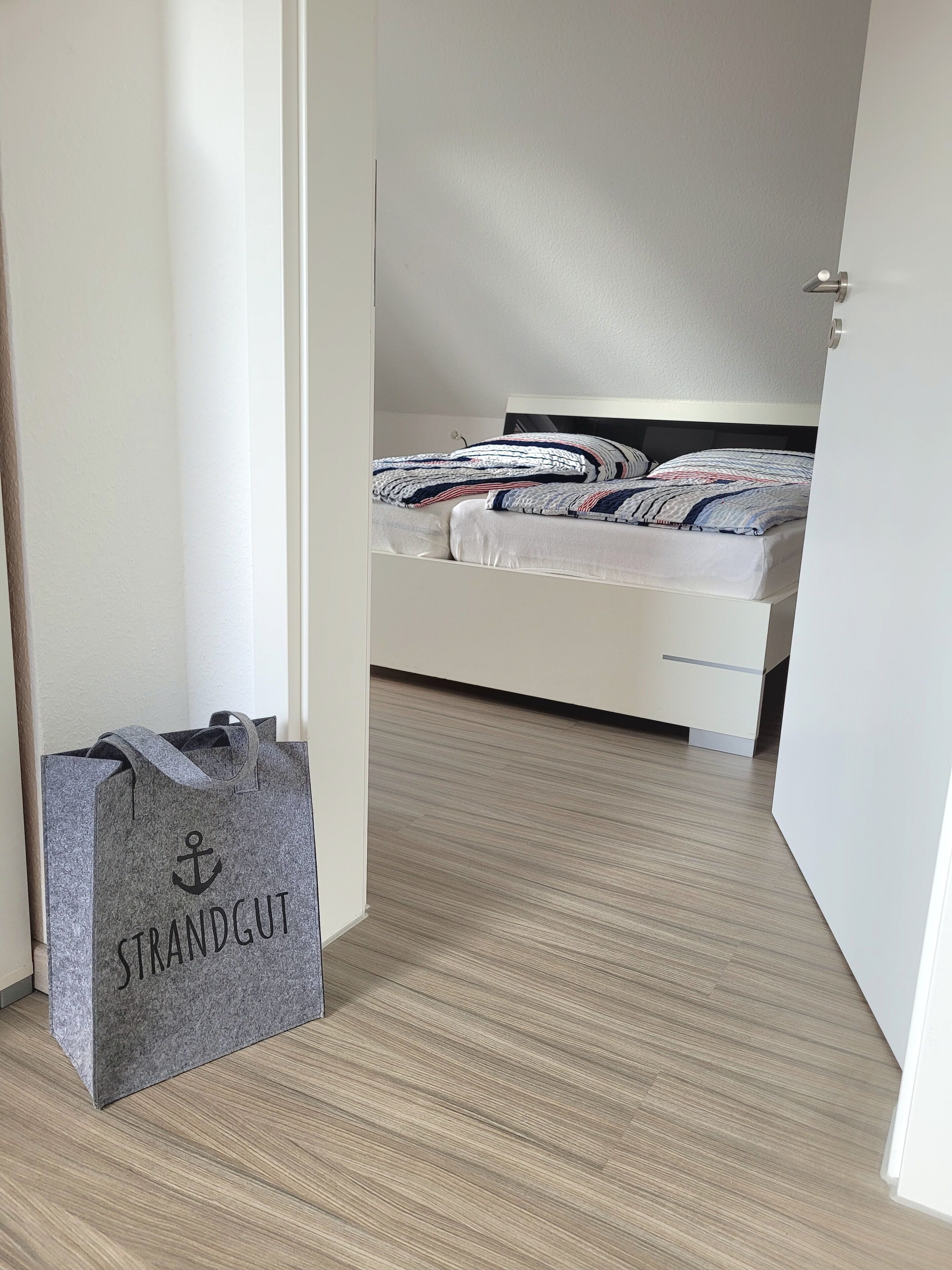 Ein Schlafzimmer mit einem Bett, einem weißen Kleiderschrank und einer grauen Tasche mit dem Aufdruck 'STRANDGUT' auf dem Boden.
