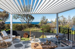 Un patio exterior con pérgola blanca, mobiliario para sentarse y vista al mar.